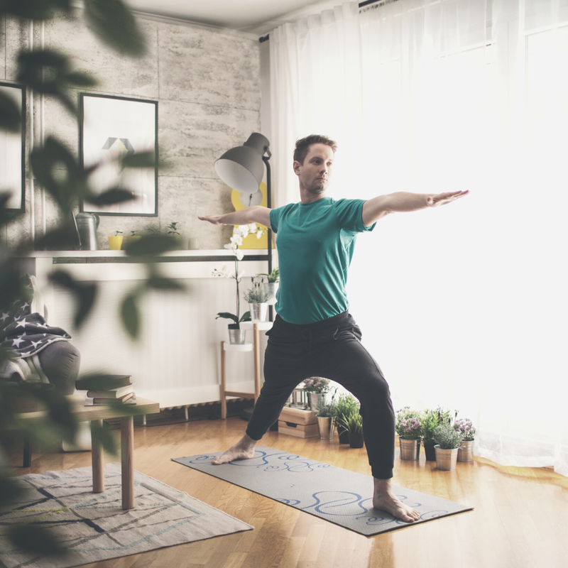 Man dressed in green t-shirt and black sweatpants practices Warrior II on a black mat in a living room with lots of plants. 