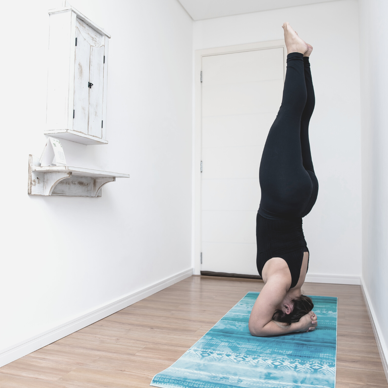 Adult dressed in all black athletic wear practices headstand in a white hallway on a blue yoga mat