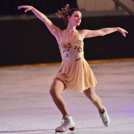 A female figure skater wearing a gold dress and white skates stops dramatically and poses under the spotlight during an ice show
