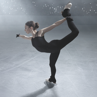 A female figure skater dressed in all black with white skates, dark hair in a bun, performs a skating element called a catch spiral. She is standing on one leg with the other leg extended to the back above the hip and is catching the lifted foot with the same hand. 