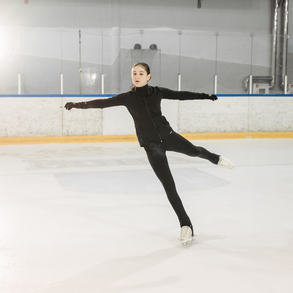 Young figure skater with black hair in a ponytail is dressed in all black with white skates. She is practicing gliding on a backward outside edge with the free leg extended