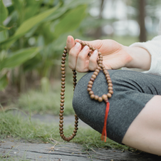 One side of a person is pictured sitting next to a green plant. They are sitting cross legged on the ground, holding a set of mala beads. 
