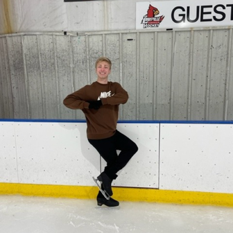 A blonde male figure skater is wearing black pants and a brown sweatshirt and black skates. He is practicing Eagle pose on the ice.  