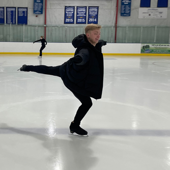 A blonde male figure skater is wearing black pants, a black hooded puffy jacket and black skates. He is executing a very strong jump landing position, with arms extended, skating leg deeply bent, free leg extended and back strong. 